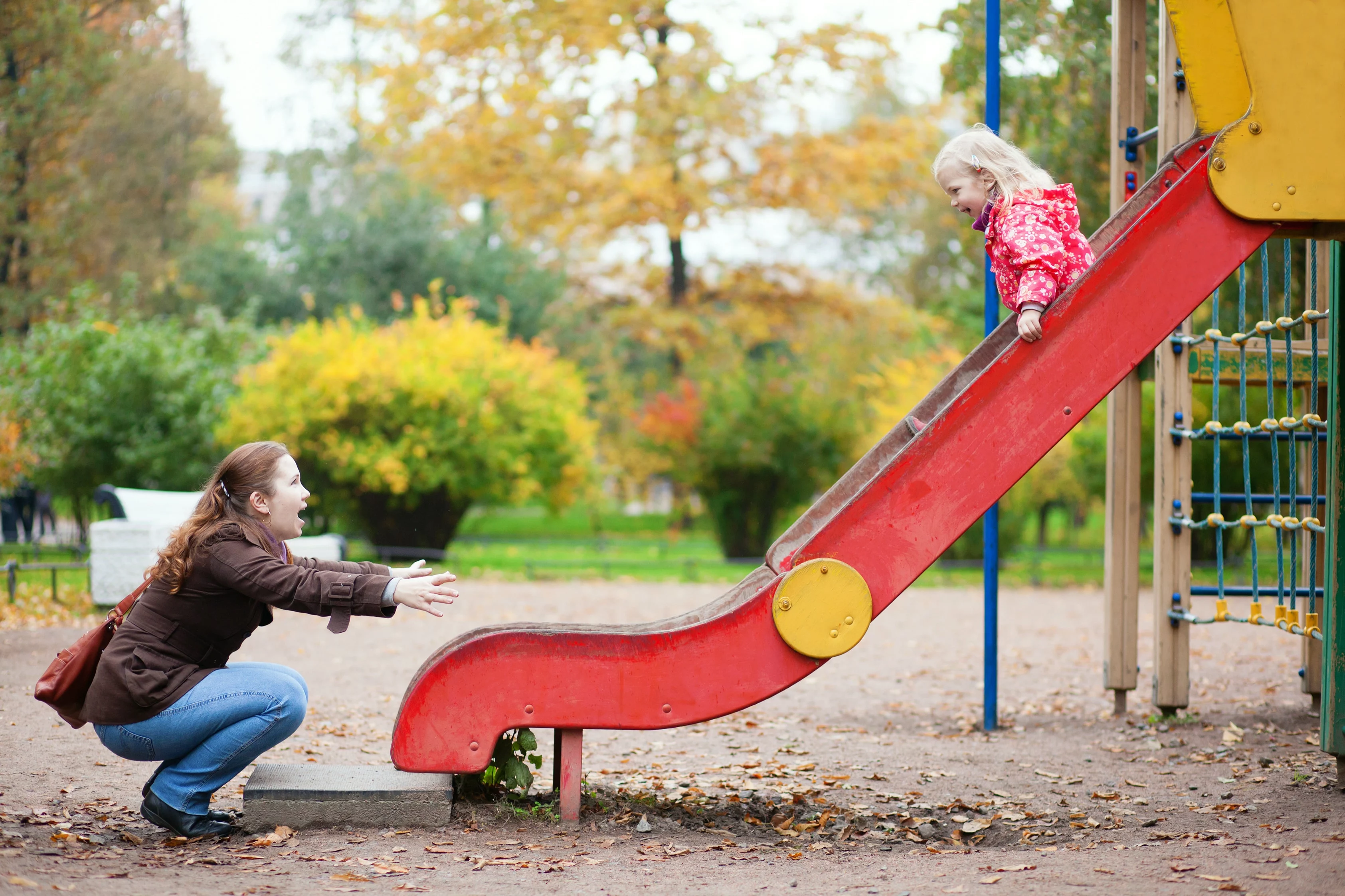 Enthousiaste moeder met kind bij een speeltuin. Kindje gaat van een rode glijbaan en lacht.